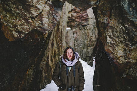 man wearing brown coat standing beside brown rock formation during daytime thumbnail