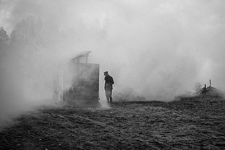 grayscale photography of a man standing next to wooden house with smokes thumbnail