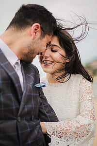 smiling woman with braces wearing white lace long-sleeved wedding dress facing smiling man wearing blue and black windowpane check double-breasted blazer thumbnail