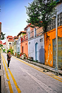 man in brown and white dress shirt standing in front of blue, orange, pink, and beige concrete building and green leaf tree thumbnail