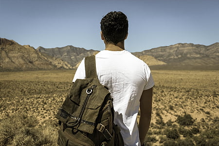 man in white shirt carrying backpack standing in front of mountain thumbnail