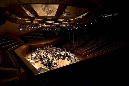 orchestra practicing on stage surrounded by benches and chairs inside building with professional lighting thumbnail