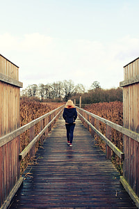 woman in black jacket and denim pants walking on wooden bridge under cloudy sky during daytime thumbnail