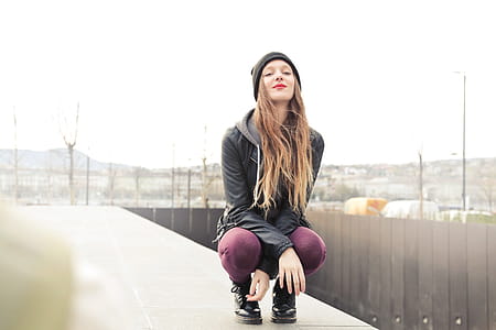 A young blonde woman wearing a black leather jacket and purple red leggings sitting on her toes in squatting posture thumbnail