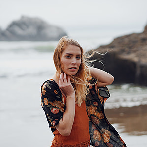 woman wearing orange spaghetti strap top with black and red floral cardigan standing on seashore with rocks during daytime thumbnail