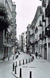 grayscale photography of people walking beside road in between buildings thumbnail