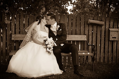 sepia photography of groom and bride kissing while sitting on bench thumbnail