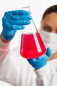 person in white laboratory gown and blue gloves holding clear glass laboratory flask with red liquid thumbnail