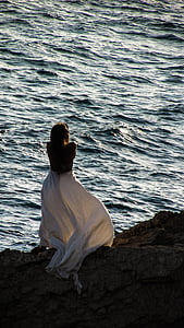 woman wearing white dress facing body of water on brown rock formation thumbnail