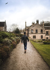 man walking on pathway beside grass field, plants and buildings at the distance during daytime thumbnail