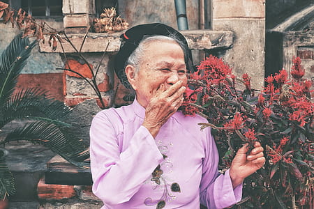 woman in pink long-sleeved shirt and black hat laughing beside red flowers and sago palm plant during daytime thumbnail