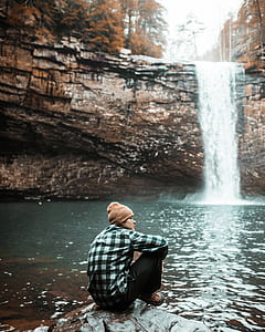 person in brown knit cap sitting on rock in front of falls thumbnail