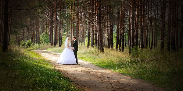 man and woman wearing wedding dress standing on road between trees thumbnail