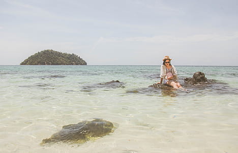 Woman Wearing White Top Sitting on Brown Rock on Body of Water during thumbnail