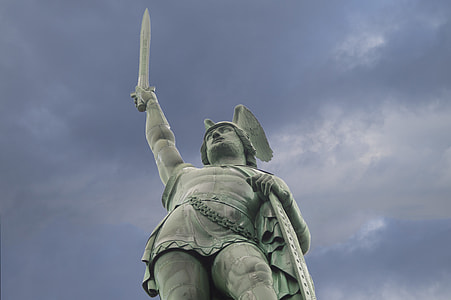 man raising sword and holding shield concrete statue under gray sky during daytime thumbnail