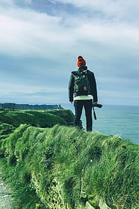 man wearing backpack holding DSLR camera standing on hill near body of water thumbnail
