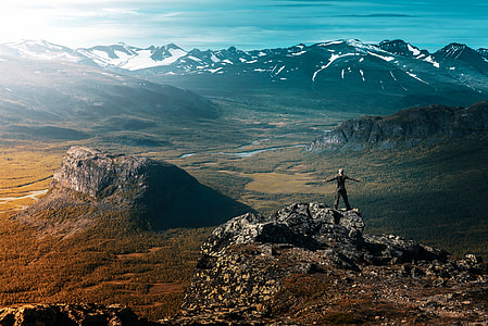 person standing on cliff facing mountains under clear blue sky thumbnail