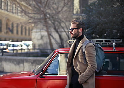 Young Adult Man in beige jacket with backpack on his shoulders,walking on the road as red vintage car seen beside him thumbnail
