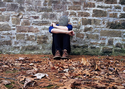 person wearing blue top and black pants sitting down near brown brick wall with head resting on arms thumbnail