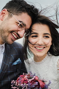 newlywed couple in wedding attire smiles and holds pink and purple flower bouquet thumbnail