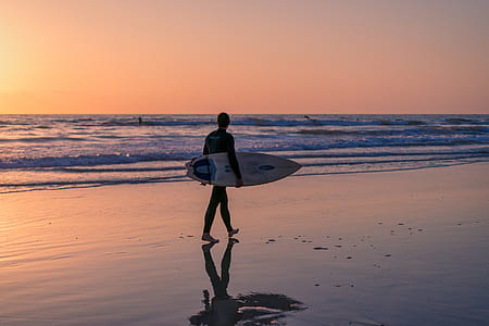 silhouette photography of man walking on beach while holding surfboard thumbnail