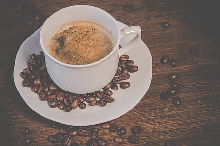 white coffee cup on saucer surrounded with coffee beans on brown wooden surface thumbnail