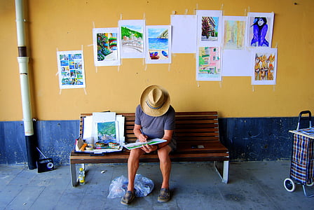 man reading book while sitting on bench near wall pasted with assorted paintings thumbnail