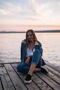 woman wearing brown denim jacket sitting on brown wood plank near water thumbnail