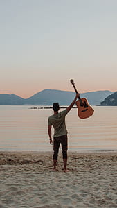 man standing on seashore holding guitar raised up during daytime thumbnail