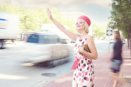 woman wearing white, pink, and brown polka-dot skater dress and red cap standing beside street waving her hand thumbnail