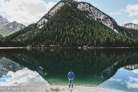 man standing in front of body of water near tree covered mountain during daytime thumbnail