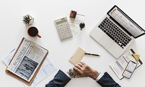 person leaning both hands on desk next to newspaper, pen, calculator, MacBook and iPhone thumbnail