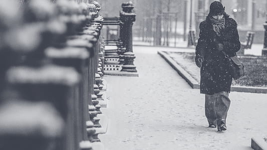 grayscale photography of a person in black trench coat walking across the street during winter weather thumbnail
