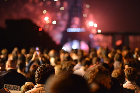 This photo displays a crowd of people who stand in front of the Eiffel Tower and who are watching the red colors of fireworks which seems to be happening behind and around the Eiffel Tower. On the lower left side a policeman is visible. thumbnail
