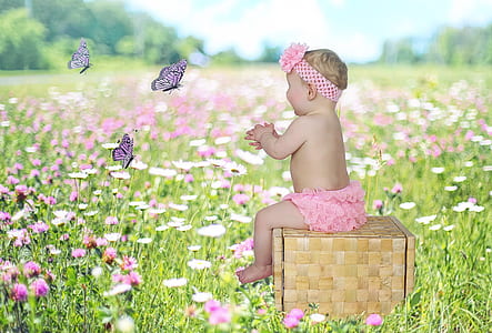 baby girl sitting on woven box in grass field surrounded by pink and white flowers thumbnail