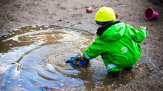 boy wearing green hooded jumpsuit playing blue toy truck on water during daytime thumbnail