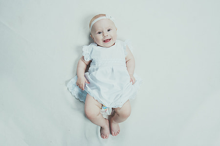 baby smiling wearing white headband and dress lying on bed thumbnail