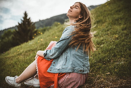 Woman Wearing Blue Denim Jacket Sitting on Green Grass Near Trees Under Blue Sky at Daytime thumbnail