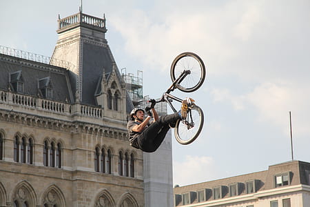 man doing bike tricks near concrete buildings under cloudy sky thumbnail