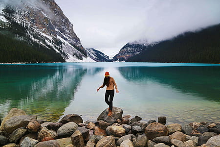 woman standing on stones beside body of water surround by mountains thumbnail