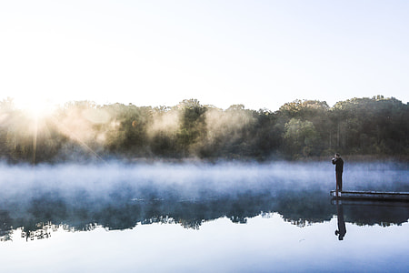 person standing on dock near calm river water surrounded by misty fogs thumbnail