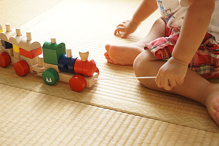 baby wearing white shirt holding assorted-color wooden train toy sitting on brown wooden flooring thumbnail
