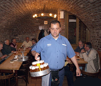 man wearing blue button-up t-shirt holding grey tray with beers inside a bar thumbnail