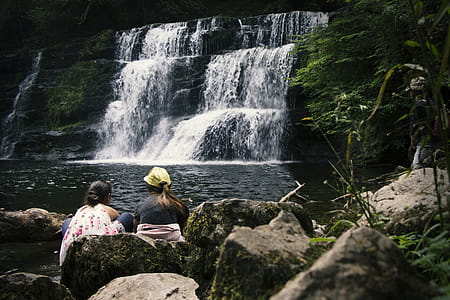 Two Women Sitting on Rock Near Waterfalls thumbnail