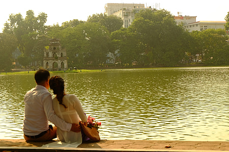 man and woman sitting on brown brick pavement looking over green body of water near trees during daytime thumbnail