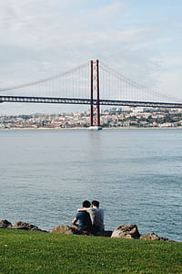 two men sitting on grass field near body of water thumbnail