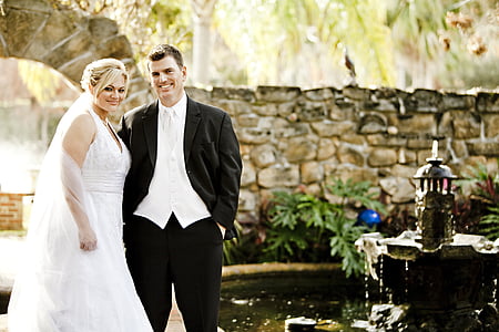 man in black suit and woman in white wedding dress standing near garden fountain during daytime thumbnail