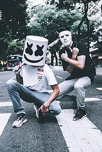 man wearing marshmallow mask holding baseball bat seating on street beside man wearing white mask thumbnail