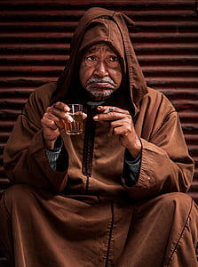 man wearing brown hooded robe holding cigarette and drinking glass while sitting thumbnail