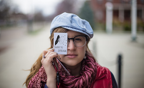 woman wearing pink and maroon zebra pattern scarf, red and black shirt, black and white plastic framed eyeglasses and blue hat while holding white Dare To Dream printed paper during daylight thumbnail